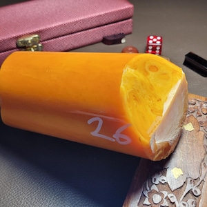 May include: A cylindrical, orange-yellow stone carving with the number 26 in white. The stone has a polished surface and a cut, angled end. A burgundy case, dice, and a carved wooden piece are in the background.