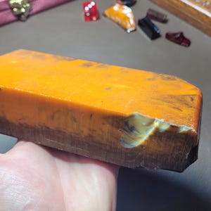 Puede incluir: Una piedra de afilar rectangular con una superficie superior de color naranja vibrante y una base de color marrón oscuro. La piedra presenta una parte superior plana y lisa con una pequeña muesca clara en la esquina frontal derecha.
