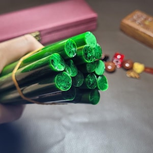 Puede incluir: Un manojo de varillas de cera cilíndricas de color verde sujetas por una banda elástica de color tostado. Las varillas tienen un exterior verde oscuro liso y una sección transversal texturizada de color verde claro en los extremos.