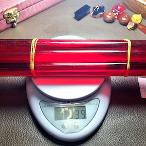 Puede incluir: Un paquete de cuatro varillas cilíndricas de color rojo translúcido sujetas con dos bandas elásticas amarillas, sobre una báscula de cocina digital blanca que muestra un peso de 39 gramos. Un pequeño papel blanco con el número 7 está al lado.