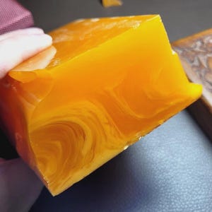 May include: A large, translucent, amber-colored block with swirling patterns is held in a hand. A maroon box and a carved wooden box are also visible. The amber block has a polished, glossy appearance.