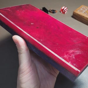 Könnte beinhalten: Ein rechteckiges Objekt aus rotem und blauem Harz mit einem weißen Streifen wird in einer Hand gehalten. Das Objekt befindet sich neben einer rosa Box und einer Holzkiste mit einem geschnitzten Design. Ein Würfelpaar und ein Ring sind ebenfalls sichtbar.