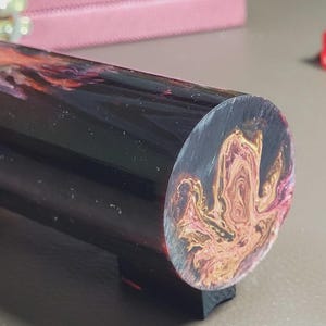 May include: A cylindrical object with a black base and a swirling pattern of orange, pink, and purple on the end. The body of the cylinder is black with a glossy finish. The object is resting on a dark surface.