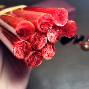 May include: A bundle of ten cylindrical red rods with a marbled, swirling texture, held together by a yellow rubber band. The rods have a glossy finish and display a deep crimson hue with lighter red streaks throughout their circular cross-sections.