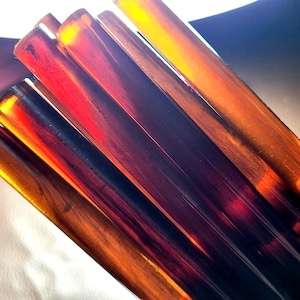 Puede incluir: Una colección de varillas cilíndricas translúcidas de color ámbar con un acabado liso y pulido. Las varillas presentan tonos variados de naranja intenso, marrón y amarillo dorado, con luz que atraviesa el material para resaltar los patrones.