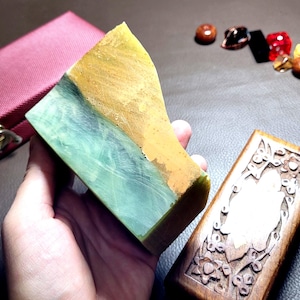 Puede incluir: Un bloque de piedra rectangular con un aspecto de dos tonos, con una sección lisa de color verde claro que transiciona a una capa superior texturizada de color amarillo mostaza. La piedra se sostiene junto a un estuche rojo y una caja de madera.