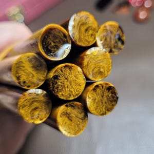 May include: A bundle of seven cylindrical rods made of a translucent amber-coloured resin with swirling black and dark brown patterns throughout. The rods are held together by a rubber band, showcasing their circular cross-sections and smooth finish.