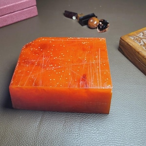 May include: An orange-red stone seal with a rounded corner, alongside a wooden box with carved floral designs. A small, maroon box and several small objects are also visible.