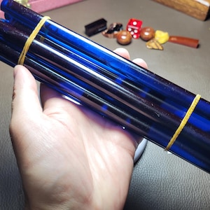 May include: A bundle of five cylindrical, translucent cobalt blue glass rods held together by two yellow rubber bands. The rods are smooth, straight, and uniform in length, featuring a deep blue hue that allows light to pass through the material.