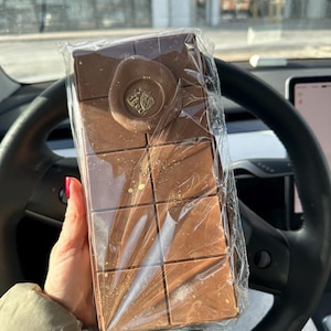May include: A rectangular chocolate bar, wrapped in clear plastic, held in a person's hand. The bar is divided into squares and has a circular emblem on top. Gold flecks are visible on the chocolate.
