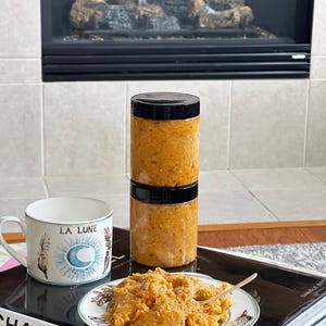 May include: Two clear plastic jars filled with orange food are stacked on top of each other. A small plate with a portion of the orange food and a spoon sits on a stack of books. A white mug with the words "LA LUNE" is also present.