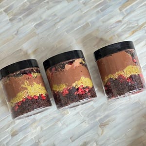 May include: Three glass jars filled with layers of chocolate cake, chocolate frosting, and red and gold sprinkles. The jars are on a white tiled surface.