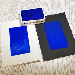 Pode incluir: Um conjunto de tintas aquarela com um tom azul vibrante rotulado como "Basket Blue". Duas amostras retangulares do mesmo azul s&atilde;o exibidas em papel branco e preto. O papel branco tem uma borda decorativa.