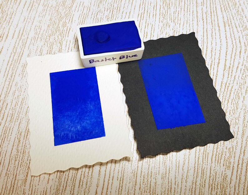 Pode incluir: Um conjunto de tintas aquarela com um tom azul vibrante rotulado como "Basket Blue". Duas amostras retangulares do mesmo azul s&atilde;o exibidas em papel branco e preto. O papel branco tem uma borda decorativa.