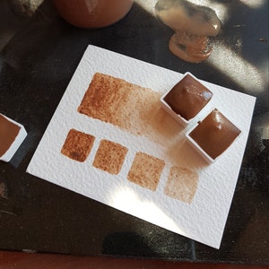 K&ouml;nnte beinhalten: Ein Aquarellfarbenset mit Braunt&ouml;nen. Das Bild zeigt ein wei&szlig;es Papier mit einer Farbmusterkarte und mehreren Quadraten mit verschiedenen Braunt&ouml;nen. Zwei Farbbeh&auml;lter sind mit dunkelbrauner Farbe gef&uuml;llt.