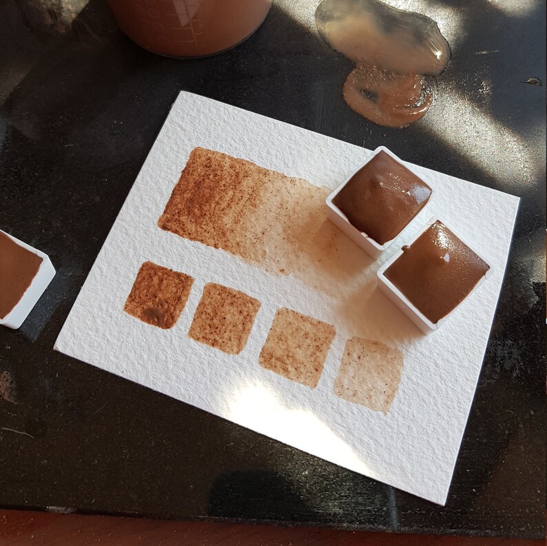 K&ouml;nnte beinhalten: Ein Aquarellfarbenset mit Braunt&ouml;nen. Das Bild zeigt ein wei&szlig;es Papier mit einer Farbmusterkarte und mehreren Quadraten mit verschiedenen Braunt&ouml;nen. Zwei Farbbeh&auml;lter sind mit dunkelbrauner Farbe gef&uuml;llt.