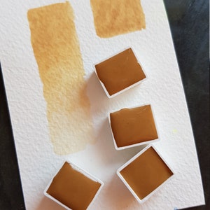 K&ouml;nnte beinhalten: Aquarellfarbenset mit vier quadratischen Farbt&ouml;pfchen brauner Farbe und einem Farbmuster der gleichen Farbe auf wei&szlig;em Papier. Das Papier hat auch zwei zus&auml;tzliche braune Farbmuster. Die Farbe hat einen warmen, erdigen Ton.