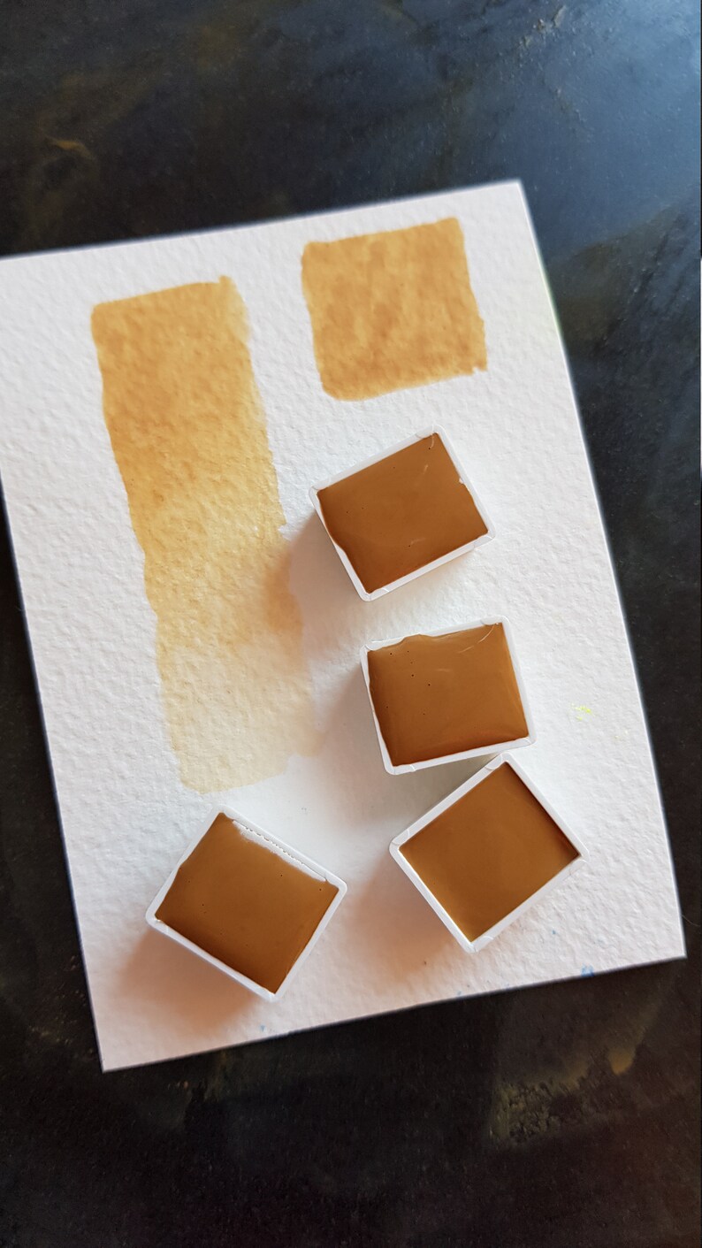 K&ouml;nnte beinhalten: Aquarellfarbenset mit vier quadratischen Farbt&ouml;pfchen brauner Farbe und einem Farbmuster der gleichen Farbe auf wei&szlig;em Papier. Das Papier hat auch zwei zus&auml;tzliche braune Farbmuster. Die Farbe hat einen warmen, erdigen Ton.