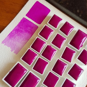May include: A collection of magenta watercolor paint pans arranged on a white surface. A swatch of the same color is painted on the paper, showing a gradient from dark to light. The paint pans are square and rectangular, filled with a glossy, vibrant pigment.