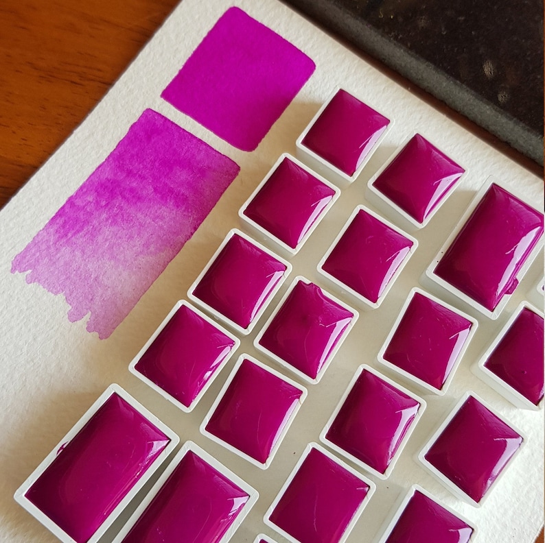 May include: A collection of magenta watercolor paint pans arranged on a white surface. A swatch of the same color is painted on the paper, showing a gradient from dark to light. The paint pans are square and rectangular, filled with a glossy, vibrant pigment.