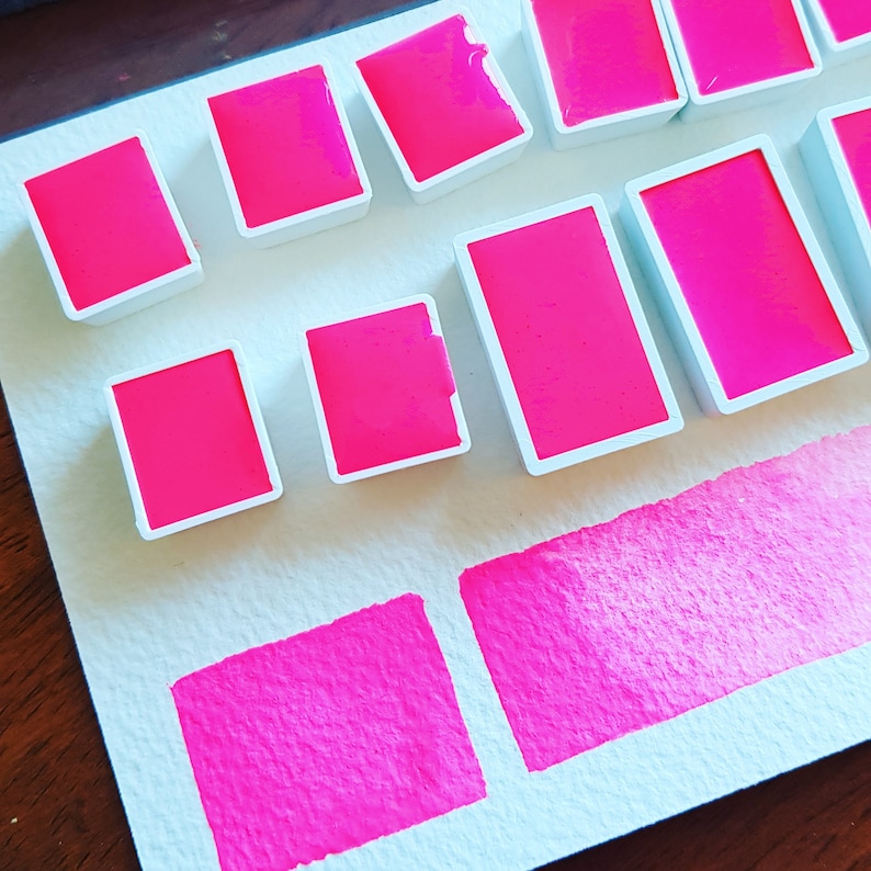 May include: Bright pink watercolor paints in rectangular pans with white borders are arranged on a white surface. Below the pans are two rectangular swatches of the same vibrant pink color, showcasing the paint's texture.