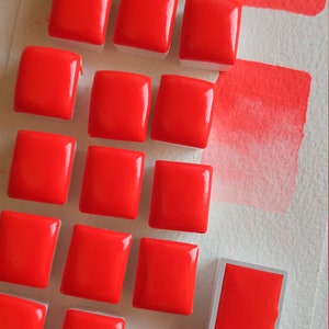 May include: A collection of vibrant red watercolor paint squares arranged on a white surface. The glossy, square paint blocks are accompanied by two swatches of the same bright red color, showcasing the pigment's intensity. Art supplies.