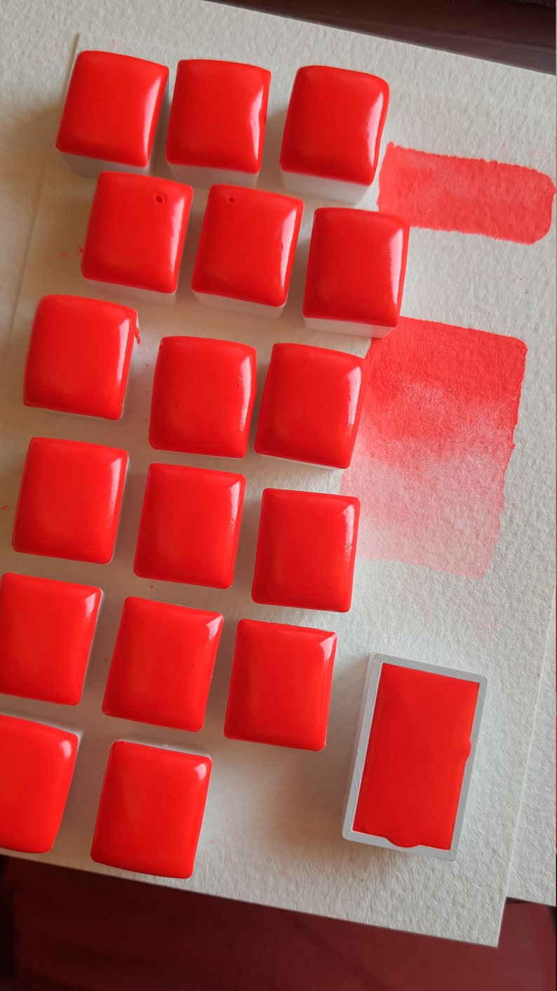 May include: A collection of vibrant red watercolor paint squares arranged on a white surface. The glossy, square paint blocks are accompanied by two swatches of the same bright red color, showcasing the pigment's intensity. Art supplies.