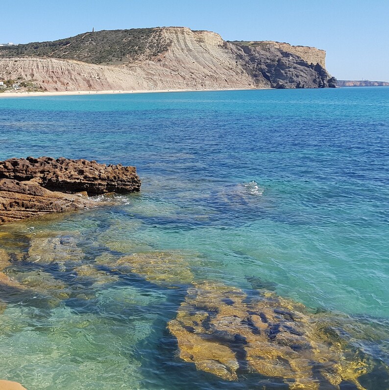 K&ouml;nnte beinhalten: Eine malerische K&uuml;stenlandschaft mit klarem t&uuml;rkisfarbenem Wasser, Felsformationen und einem Sandstrand. Im Hintergrund erhebt sich eine gro&szlig;e Klippe mit geschichteten Felsformationen unter einem klaren blauen Himmel. Das Wasser spiegelt die Farbe des Himmels wider.