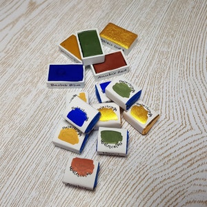 Pode incluir: Uma cole&ccedil;&atilde;o de pastilhas de aquarela em v&aacute;rias cores, incluindo azul, verde, dourado e vermelho. Cada pastilha &eacute; rotulada com um nome de cor, como "Basket Blue" e "Osiris Gold". As tintas est&atilde;o dispostas sobre uma superf&iacute;cie clara e texturizada.