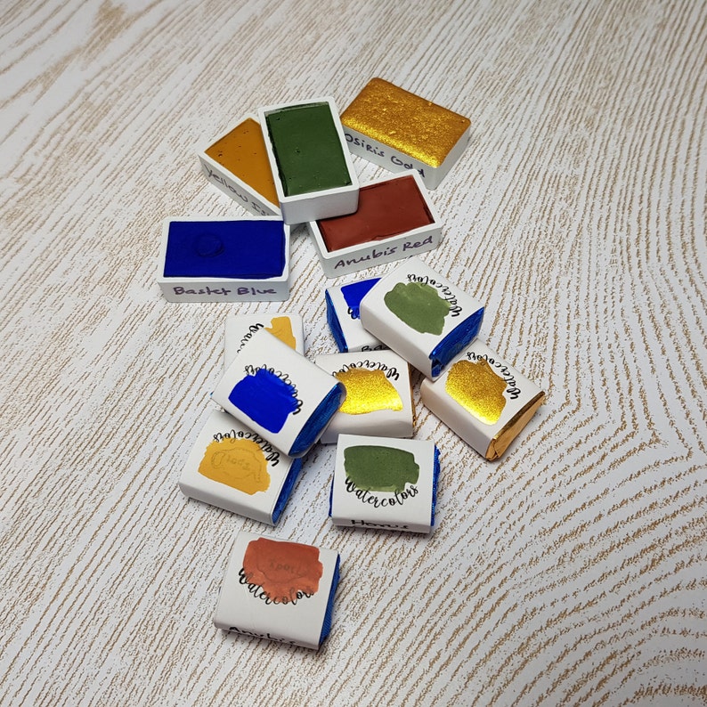 Pode incluir: Uma cole&ccedil;&atilde;o de pastilhas de aquarela em v&aacute;rias cores, incluindo azul, verde, dourado e vermelho. Cada pastilha &eacute; rotulada com um nome de cor, como "Basket Blue" e "Osiris Gold". As tintas est&atilde;o dispostas sobre uma superf&iacute;cie clara e texturizada.