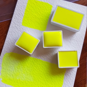 May include: A close-up of watercolor paints and swatches in a vibrant neon yellow hue. The image shows four rectangular paint pans and two painted squares on textured white paper, showcasing the color's intensity.