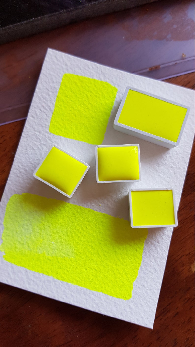 May include: A close-up of watercolor paints and swatches in a vibrant neon yellow hue. The image shows four rectangular paint pans and two painted squares on textured white paper, showcasing the color's intensity.