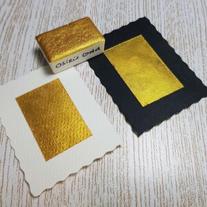 Pode incluir: Uma tinta aquarela dourada, rotulada como "Osiris Gold", est&aacute; sobre duas amostras de tinta dourada em papel branco e preto. A tinta dourada tem um brilho met&aacute;lico e o papel tem uma superf&iacute;cie texturizada.