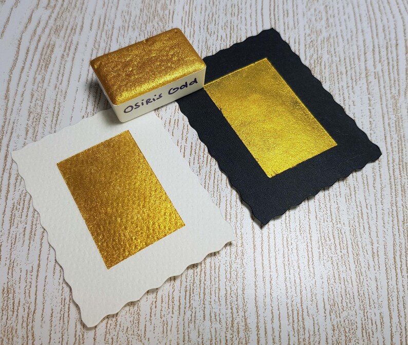 Pode incluir: Uma tinta aquarela dourada, rotulada como "Osiris Gold", est&aacute; sobre duas amostras de tinta dourada em papel branco e preto. A tinta dourada tem um brilho met&aacute;lico e o papel tem uma superf&iacute;cie texturizada.