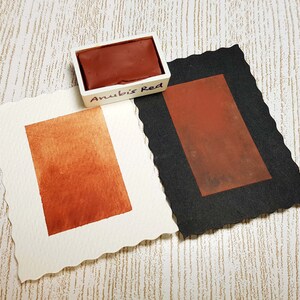 Pode incluir: Um conjunto de tintas aquarela rotulado "Anubis Red" com um tom marrom-avermelhado profundo. Duas amostras de papel aquarela exibem a cor, uma em papel branco e outra em papel preto. O papel branco tem uma borda irregular.