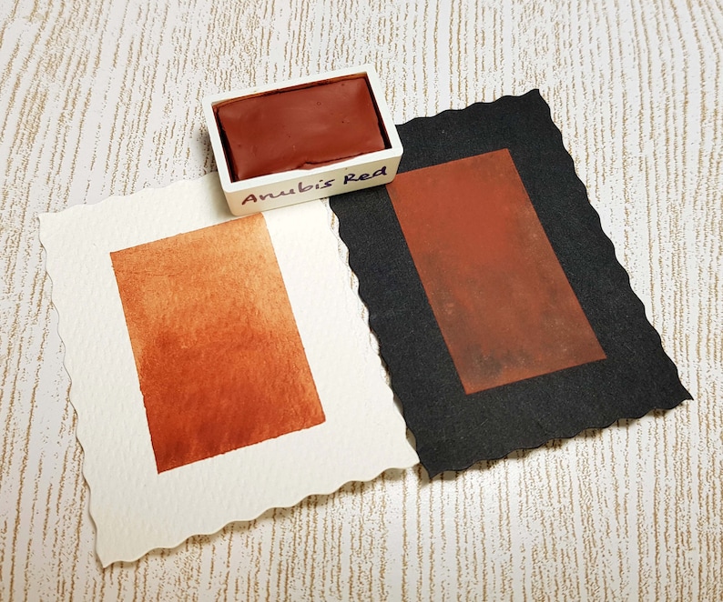 Pode incluir: Um conjunto de tintas aquarela rotulado "Anubis Red" com um tom marrom-avermelhado profundo. Duas amostras de papel aquarela exibem a cor, uma em papel branco e outra em papel preto. O papel branco tem uma borda irregular.