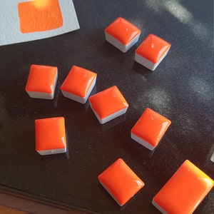 May include: Bright orange watercolor paint pans with a glossy finish, arranged on a dark surface. A swatch of the same orange color is on a piece of white paper in the background. The paint pans have a white base.