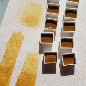 K&ouml;nnte beinhalten: Aquarellfarbenset mit mehreren kleinen, wei&szlig;en, rechteckigen Beh&auml;ltern, die mit einem satten, braunen Pigment gef&uuml;llt sind. Auf wei&szlig;em Papier sind Farbmuster in der gleichen Farbe aufgetragen, die die Transparenz und den Farbton der Farbe demonstrieren. Die Kunstutensilien sind auf einer wei&szlig;en Oberfl&auml;che angeordnet.
