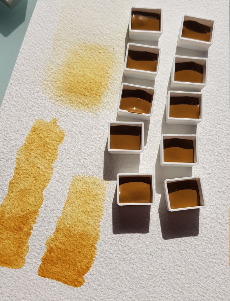 K&ouml;nnte beinhalten: Aquarellfarbenset mit mehreren kleinen, wei&szlig;en, rechteckigen Beh&auml;ltern, die mit einem satten, braunen Pigment gef&uuml;llt sind. Auf wei&szlig;em Papier sind Farbmuster in der gleichen Farbe aufgetragen, die die Transparenz und den Farbton der Farbe demonstrieren. Die Kunstutensilien sind auf einer wei&szlig;en Oberfl&auml;che angeordnet.