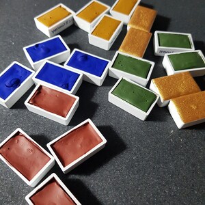 Pode incluir: Uma cole&ccedil;&atilde;o de pastilhas de tinta aquarela em v&aacute;rias cores, incluindo amarelo, azul, verde, marrom e dourado. Cada pastilha &eacute; retangular e est&aacute; em um recipiente de pl&aacute;stico branco. Algumas pastilhas t&ecirc;m etiquetas manuscritas.