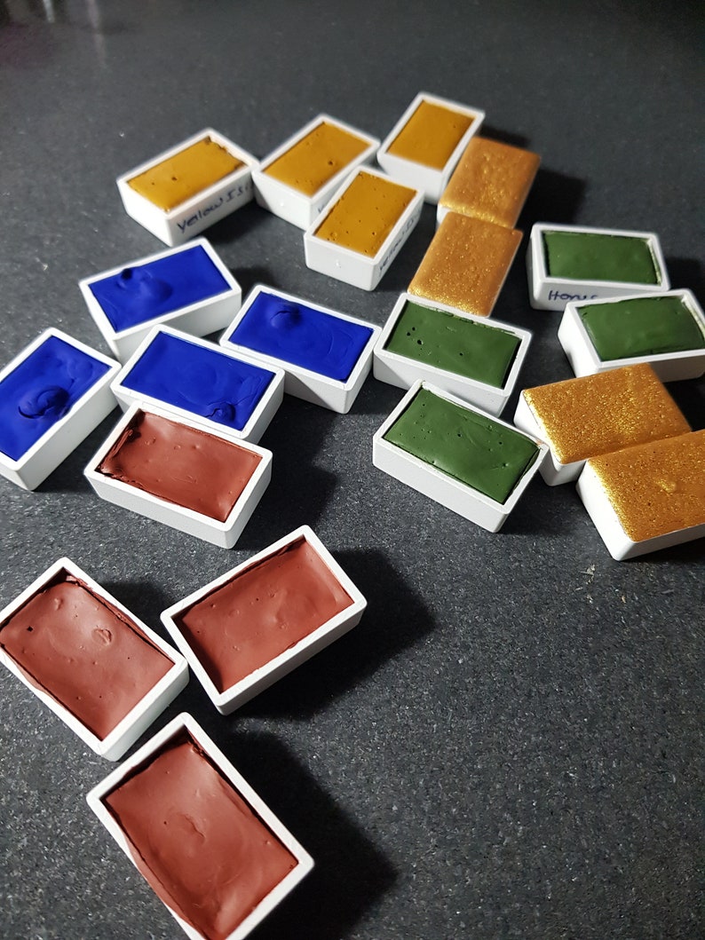 Pode incluir: Uma cole&ccedil;&atilde;o de pastilhas de tinta aquarela em v&aacute;rias cores, incluindo amarelo, azul, verde, marrom e dourado. Cada pastilha &eacute; retangular e est&aacute; em um recipiente de pl&aacute;stico branco. Algumas pastilhas t&ecirc;m etiquetas manuscritas.