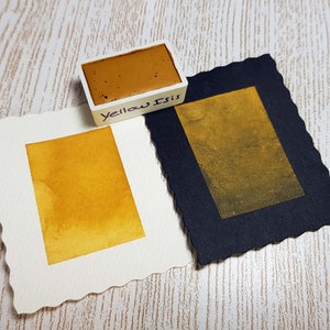 Pode incluir: Um conjunto de tintas aquarela com um bloco de tinta amarelo-marrom rotulado "Yellow Isis" ao lado de duas amostras de aquarela. Uma amostra est&aacute; em papel branco, a outra em papel preto, ambas exibindo um tom amarelo dourado.