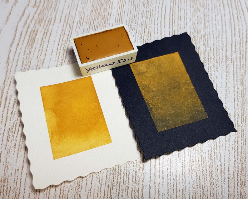 Pode incluir: Um conjunto de tintas aquarela com um bloco de tinta amarelo-marrom rotulado "Yellow Isis" ao lado de duas amostras de aquarela. Uma amostra est&aacute; em papel branco, a outra em papel preto, ambas exibindo um tom amarelo dourado.