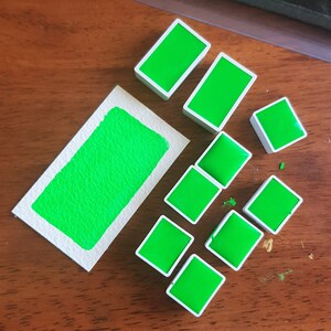 May include: A collection of vibrant green watercolor paint blocks and a swatch on a piece of paper. The paint blocks are rectangular with white borders. The swatch shows the texture of the paint.