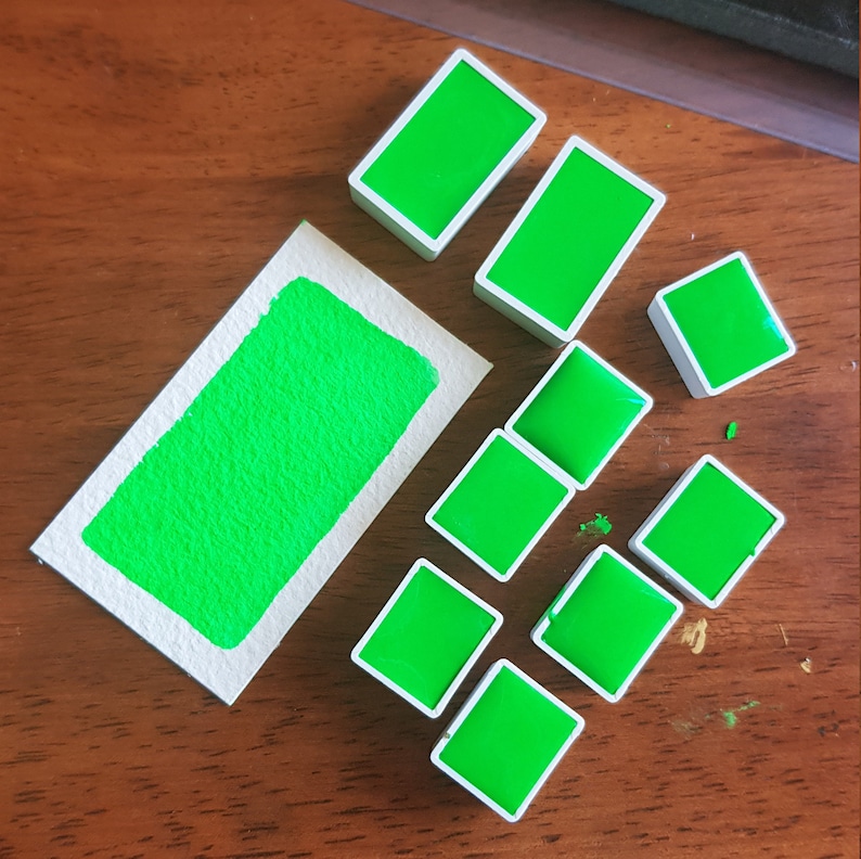 May include: A collection of vibrant green watercolor paint blocks and a swatch on a piece of paper. The paint blocks are rectangular with white borders. The swatch shows the texture of the paint.
