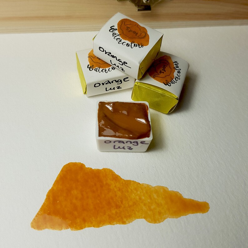 K&ouml;nnte beinhalten: Orange Aquarellfarben, einschlie&szlig;lich einer Farbprobe. Die Farbe befindet sich in kleinen, quadratischen, wei&szlig;en Beh&auml;ltern mit der Aufschrift "Orange Luz". Die Beh&auml;lter sind in gelbes und wei&szlig;es Papier gewickelt.