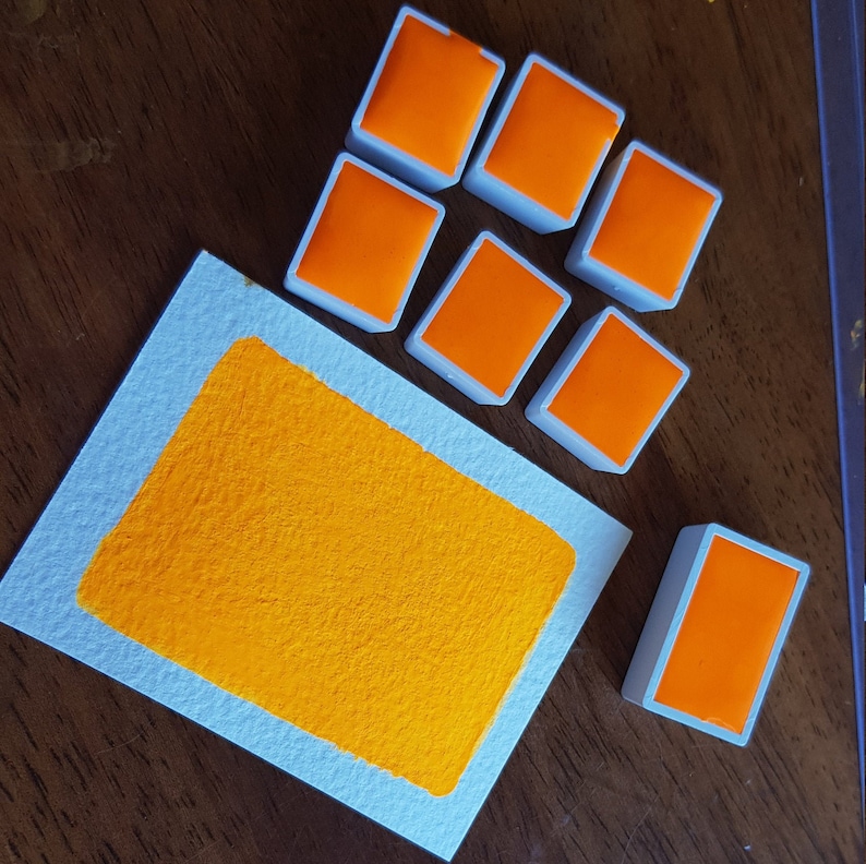May include: A collection of orange watercolor paint pans and a swatch of orange paint on a white paper. The paint pans are rectangular with a white border. The paper has a light blue border.