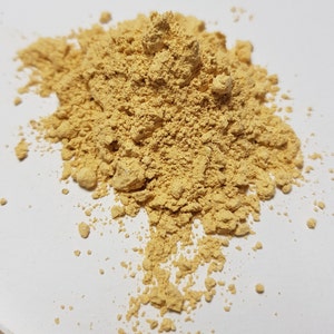 May include: A close-up shot of a pile of light brown powder against a white background. The powder has a fine, granular texture and appears to be a natural substance. The lighting is bright and even, highlighting the texture of the powder.