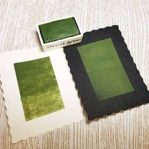 Pode incluir: Um conjunto de tintas aquarela com um bloco de tinta "Horus Green" em um recipiente branco. Duas amostras de papel aquarela exibem o mesmo tom verde, uma em papel branco e outra em papel preto. As bordas do papel s&atilde;o recortadas.