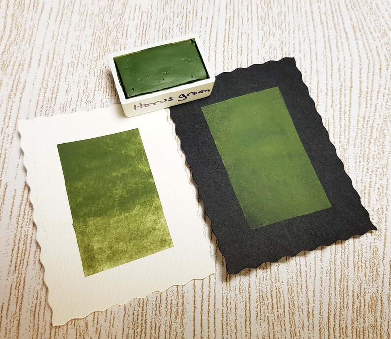 Pode incluir: Um conjunto de tintas aquarela com um bloco de tinta "Horus Green" em um recipiente branco. Duas amostras de papel aquarela exibem o mesmo tom verde, uma em papel branco e outra em papel preto. As bordas do papel s&atilde;o recortadas.